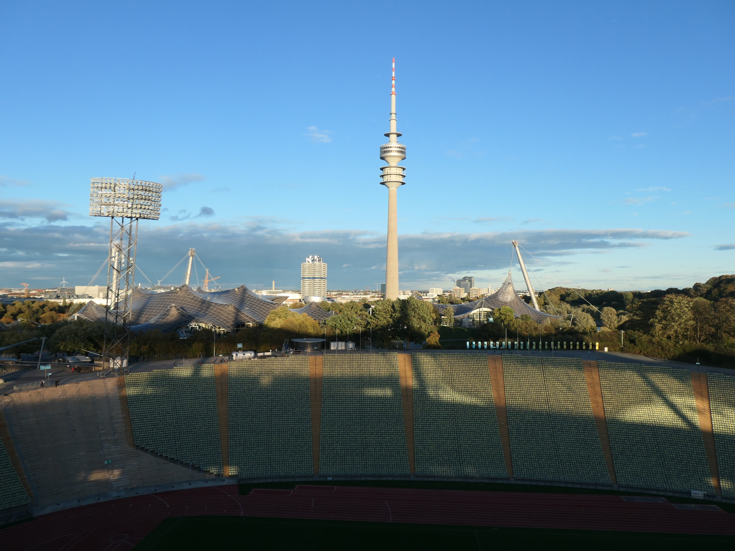 Das Olympische Stadion in Berlin, Deutschland, mit dem Fernsehturm im Hintergrund, umgeben von Bäumen, Gebäuden und beleuchteten Bereichen unter einem bewölkten Himmel.