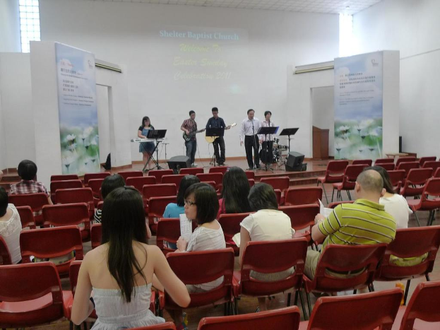 Ein Zuschauerraum mit einer Bühne, auf der zwei Personen in der Mitte Gitarren spielen und andere in den Ecken singen und Instrumente spielen, mit Schildern neben der Bühne und einem Publikum davor, das Papiere und Bücher hält.