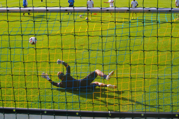 Ein Fussballer taucht ab, um einen Ball vor einem Tor zu halten, mit einem Netz im Vordergrund und einigen Menschen im Hintergrund, während mehrere Fussbälle auf dem Boden verstreut sind.