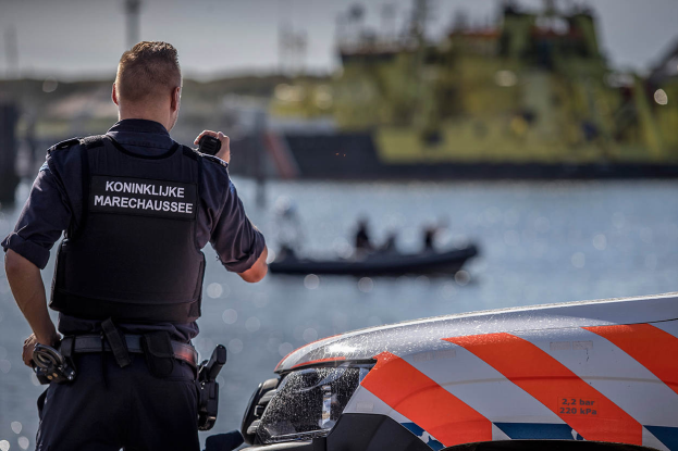 Ein Polizist in Uniform steht neben einem Polizeiwagen an einem Gewässer und hält ein Funkgerät in der Hand, im Hintergrund ist ein Boot mit ein paar Menschen auf dem Wasser zu sehen und der Himmel darüber.