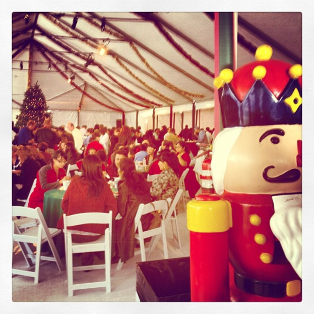 Eine Gruppe von Menschen feiert Weihnachten in einer Hütte, mit einem geschmückten Baum, weißen Stühlen, einem roten Spielzeug-Sicherheitswächter und einer schwarzen Krone.