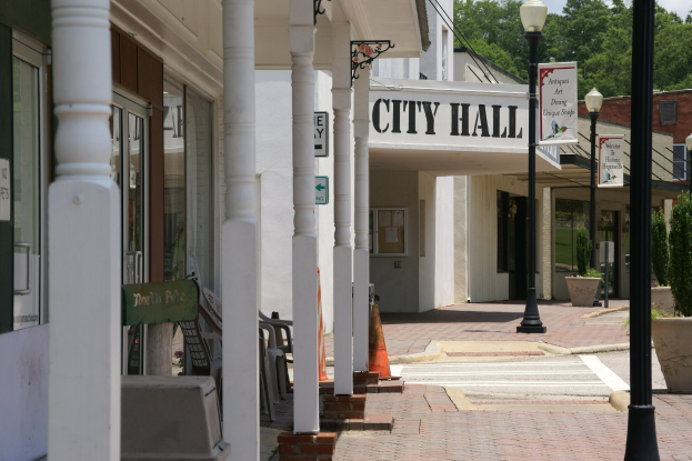Stadthallengebäude mit umgebender Infrastruktur, einschließlich Straßenlaternen, Schildern, Fahrzeugen, Verkehrskegeln, Stühlen, Topfpflanzen, Bäumen und einer bewölkten Himmel.