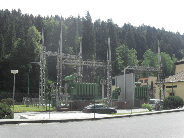Elektrische Umspannstation mit einem vorbeifahrenden Auto, umgeben von einem Zaun, Masten, Lichtern, Pflanzen, Gras und Bäumen, mit Gebäuden und einem klaren blauen Himmel im Hintergrund.