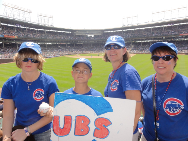 Drei Frauen in Mützen und Brillen, ein Junge in Brille und Mütze, der einen Ball und ein Schild hält, stehen in einem Stadion mit Publikum und Feld dahinter, unter einem Himmel mit ein paar Lichtern.