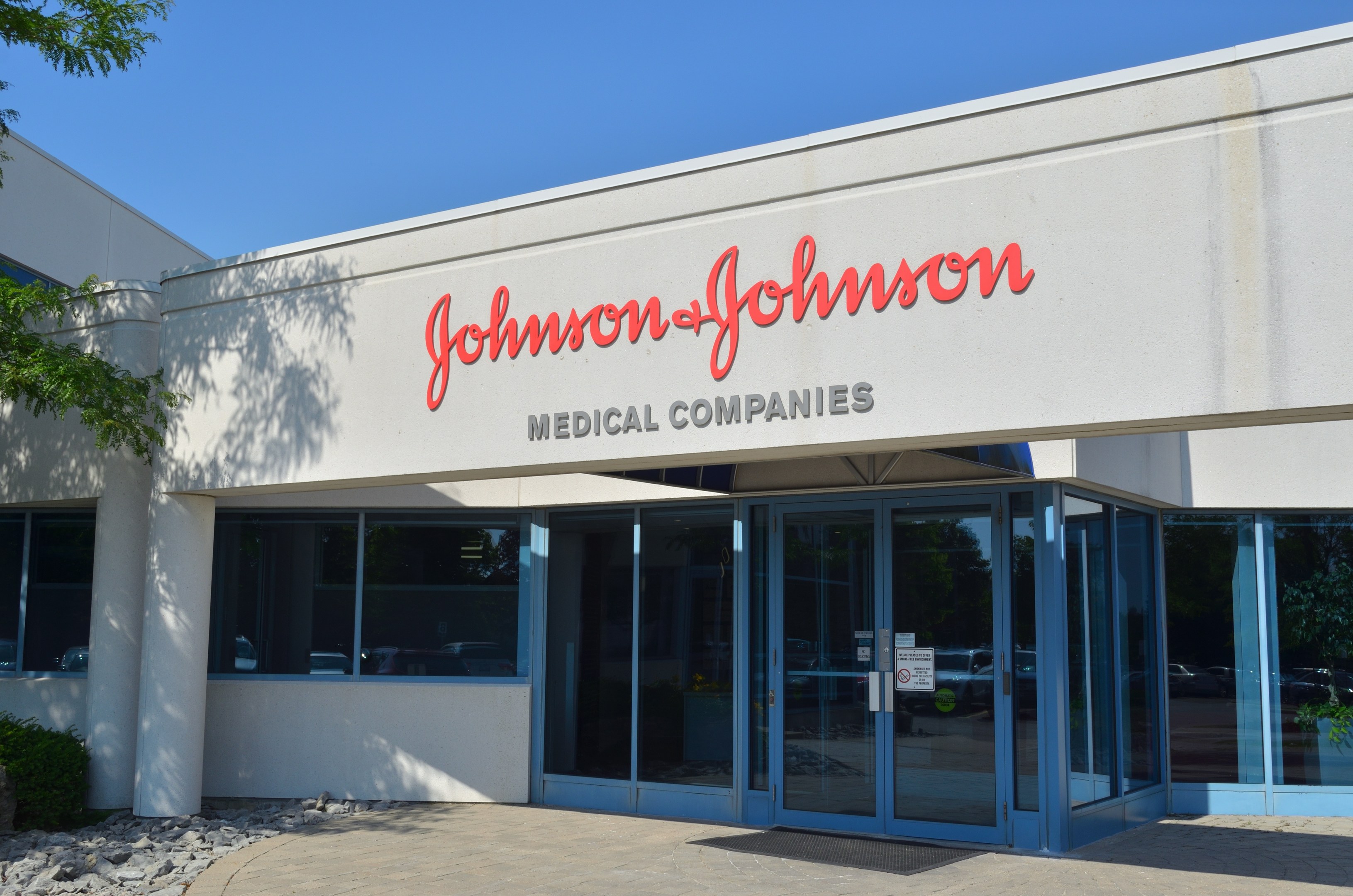 Außenansicht des Johnson & Johnson Medical Companies-Gebäudes mit Glaswänden und -türen, Wandtext, Steinen, Pflanzen und einem Baum auf der linken Seite unter einem klaren blauen Himmel.
