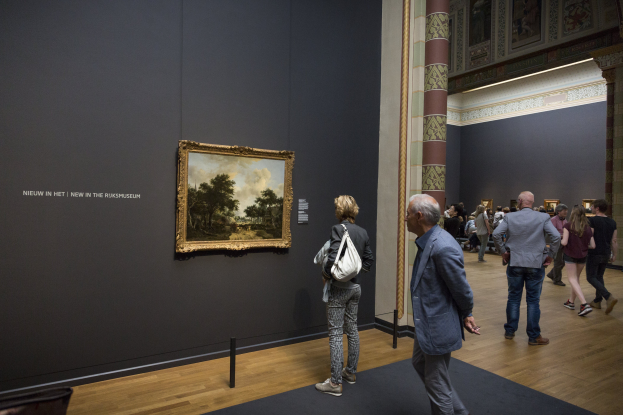 Eine Gruppe von Menschen mit Taschen geht durch ein Museum und bewundert ein gerahmtes Gemälde an der Wand mit Schutzseilen davor.