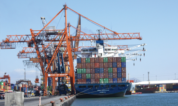 Ein Hafenbild mit großen Kränen, Waren auf einer Brücke neben dem Meer und Schiffen im Hintergrund.