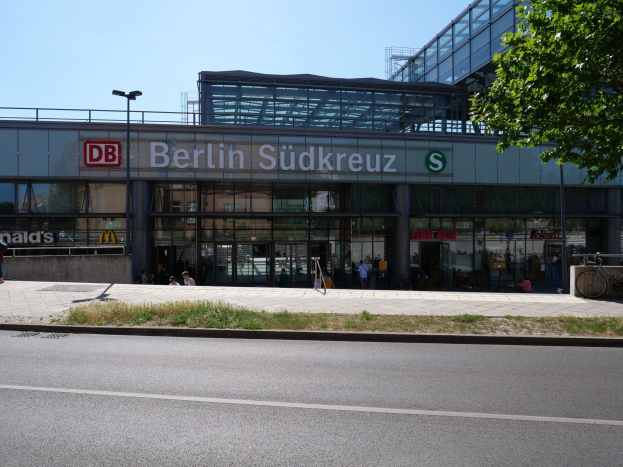 Großes gläsernes Berliner Südkreuz-Bahnhofgebäude mit Text an seiner Fassade, umgeben von Laternenmasten, Lampen, Fahrzeugen, Fußgängern, Fahrrädern, Bäumen und einem klaren blauen Himmel.