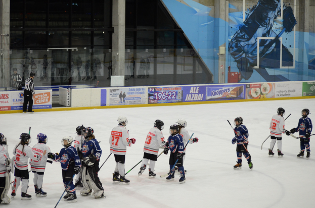 Junge Menschen in Hockey-Ausrüstung spielen Eis-Hockey auf einer Indoor-Eisfläche mit Bannern, Säulen und Glaswänden im Hintergrund.