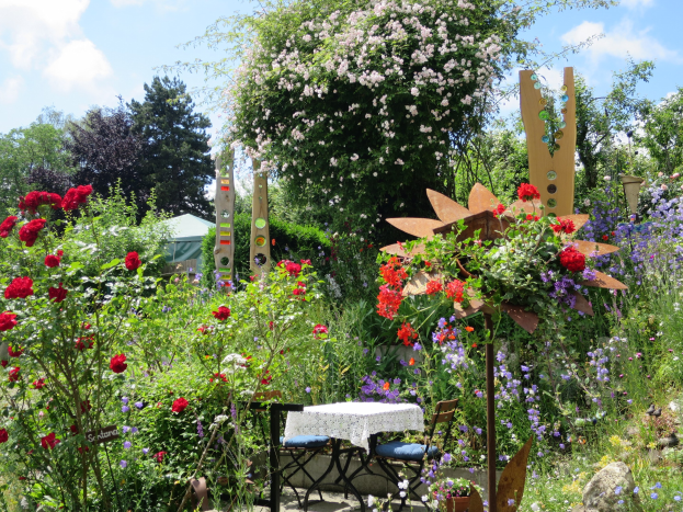 Ein Garten auf der Chelsea Flower Show mit bunten Blumen und Pflanzen, einem Tisch und Stühlen in der Mitte mit einer Decke darauf, umgeben von Bäumen unter einem bewölkten Himmel.