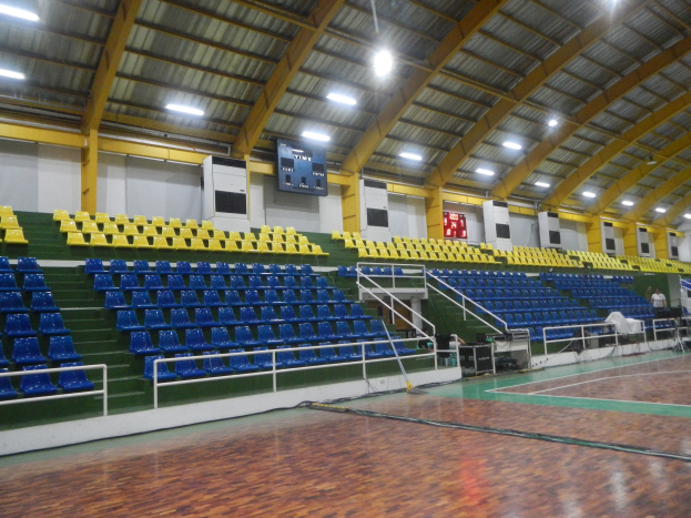 Indoor-Sporthalle mit Holzfußboden, blauen und gelben Sitzplätzen, Geländern, Treppen, wandmontierten Bildschirmen, Deckenleuchten und einigen Menschen.