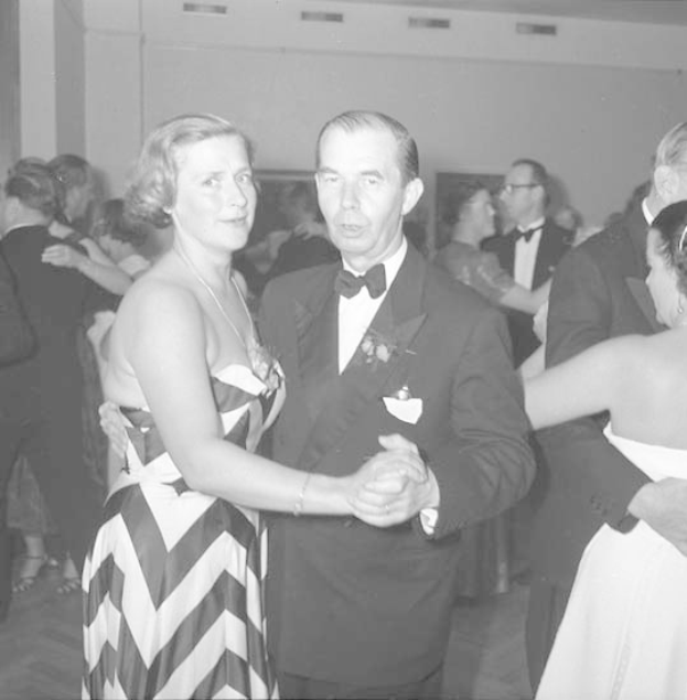 Ein Mann in einem schwarzen Blazer, weißem Hemd und schwarzen Hosen tanzt mit einer Frau in einem unbekannten Outfit auf einer formellen Veranstaltung, indem sie sich an den Händen halten und zur Musik in der Nähe einer Wand wiegen.