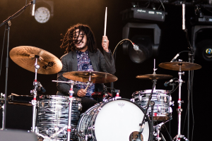 Ein Mann mit Dreadlocks spielt auf der Bühne Schlagzeug bei einem Konzert, hält Drumsticks und hat Mikrofonstative vor sich.