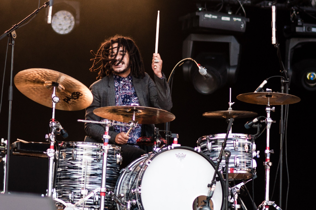 Ein Mann mit Dreadlocks spielt auf der Bühne Schlagzeug bei einem Konzert, hält Drumsticks und hat Mikrofonstative vor sich.
