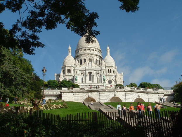 Ein weißes Gebäude umgeben von einem Zaun, Gras, Bäumen und Menschen mit Stangen und verschiedenen Pflanzen.