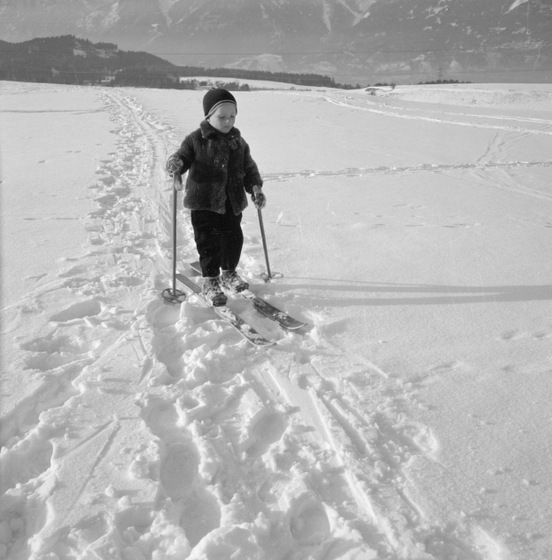 Schwarzes Bild eines jungen Jungen, der einen verschneiten Hügel mit Bergen im Hintergrund auf Skiern hinunterfährt, trägt eine Jacke, eine Mütze, Handschuhe und Schuhe und hält Ski-Stöcke in den Händen.
