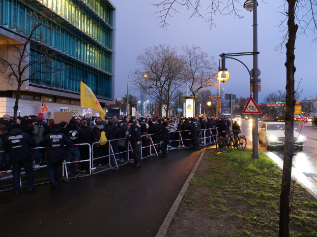 Eine große Gruppe von Menschen mit Schildern steht vor einem Gebäude mit Barrikaden, Fahrrädern, Laternenmasten, Schildern, Bäumen und Gras, mit dem Himmel im Hintergrund, was auf eine Protestaktion in Berlin hinweist.