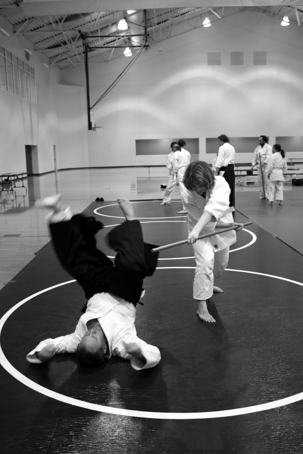 Zwei Personen üben Karate mit Stöcken in einer Turnhalle, während andere in der Nähe stehen, mit sichtbaren Lichtern an der Wand im Hintergrund.