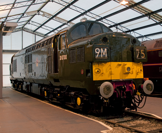 Ein Zug fährt auf den Schienen neben einer Plattform mit einem Mast auf dem Dach, der Metallstangen mit Lampen trägt.