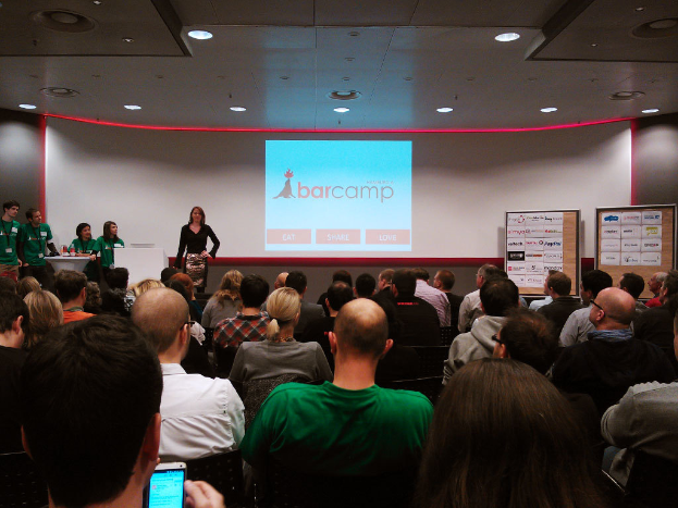 Eine Gruppe von Menschen, die auf Stühlen sitzen und einer Frau zuhören, die an einem Rednerpult spricht, mit einem Projektionsschirm, zwei Anschlagtafeln, einem Tisch und mehreren an der Decke befestigten Lampen im Hintergrund.