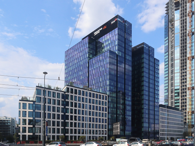 Zentrale des Deutschen Bank in Frankfurt, Deutschland, ein großes mehrstöckiges Gebäude mit zahlreichen Fenstern, umgeben von städtischer Infrastruktur einschließlich Straßenlaternen, Fahrzeugen, Grünflächen und einem bewölkten Himmel.