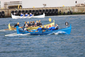 Eine Gruppe von Menschen in einem blauen und gelben Boot auf dem Wasser, die Paddel halten, mit einer Wand aus Reifen und einem Gebäude im Hintergrund.