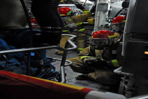 Feuerwehrleute in Helmen und Schutzausrüstung arbeiten an einem Löschfahrzeug mit Rohren und Gegenständen im Hintergrund und einer Lampe oben.
