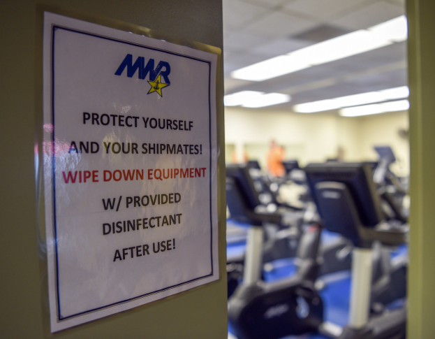 Ein Türschild mit der Aufschrift "Schützen Sie sich und Ihre Schiffskameraden" mit Laufbändern und Deckenbeleuchtung im Hintergrund.