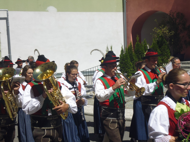 Eine Gruppe von Menschen in traditioneller bayrischer Tracht spielt Posaunen vor einem Gebäude, umgeben von Pflanzen, Geländern und einer Wand, wobei einige eine Brille tragen und Musikinstrumente oder Blumen halten.