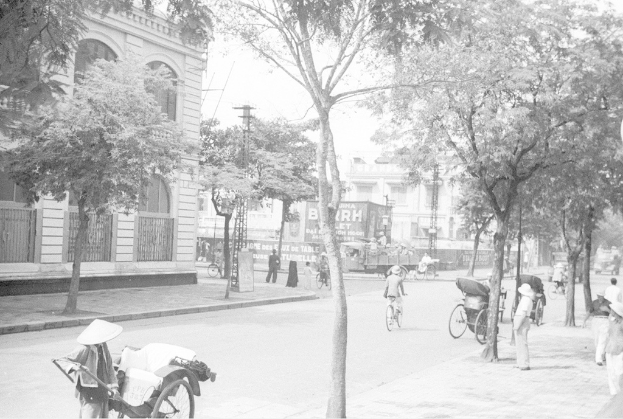 Ein altes Gebäude mit einem Fenster, hinter dem weiße Gebäude und Bäume zu sehen sind, mit drei Fahrrädern auf der rechten Seite in der Nähe eines Fußwegs und mehr Bäumen.