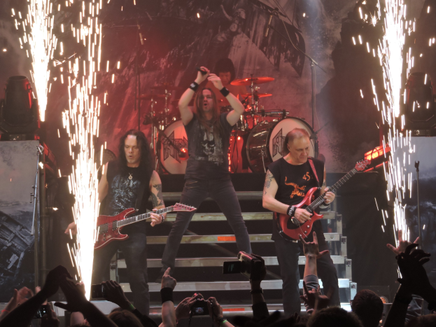 Eine Gruppe von Menschen, die auf der Bühne eines Konzerts spielen, wobei einige Gitarren halten und einer ein Mikrofon hält; Lautsprecher sind links sichtbar, und eine Menge mit Handys ist unten.