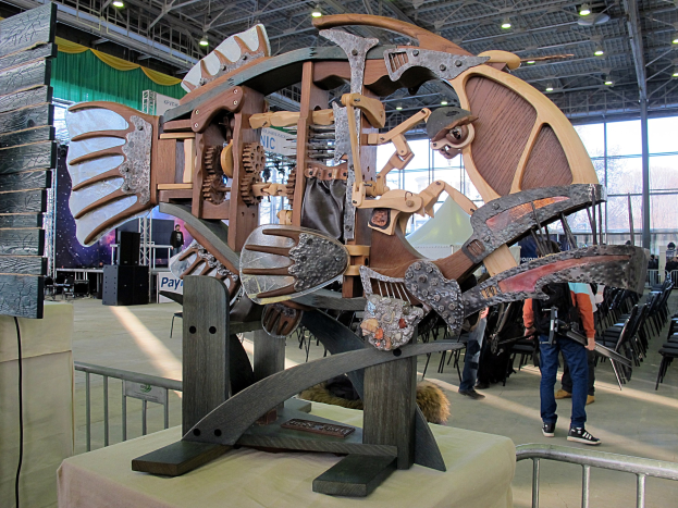 Große Metallfischskulptur in einem Raum mit Publikum, umgeben von Stühlen, einem Zaun, einer Tafel, einem Lautsprecher, Vorhängen und Deckenlampen.