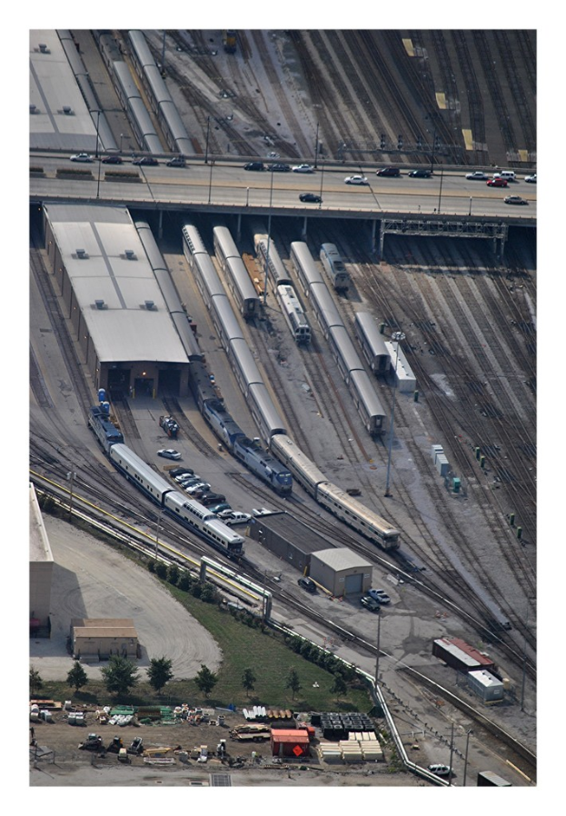 Eine Brücke mit einigen Fahrzeugen darauf, darunter Eisenbahnschienen mit einigen Zügen und eine Landschaft mit Gebäuden, Bäumen, Pfählen und diversen Gegenständen und Fahrzeugen auf dem Boden.