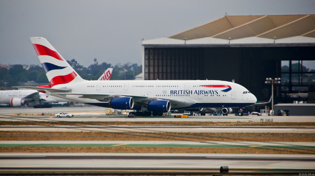 Ein Flugzeug steht auf der Rollbahn mit Straßenlaternen rechts, einem Auto in der Nähe und einem Schuppen im Hintergrund. In der Ferne gibt es Bäume, Gebäude und ein weiteres Flugzeug, das in einem offenen Bereich geparkt ist, mit einem bewölkten Himmel darüber.