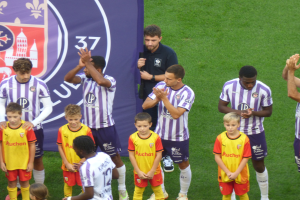 Eine Gruppe junger Jungs steht auf einem Fußballfeld, klatscht in die Hände, mit einem Banner und Logos im Hintergrund und grünem Gras unter den Füßen.