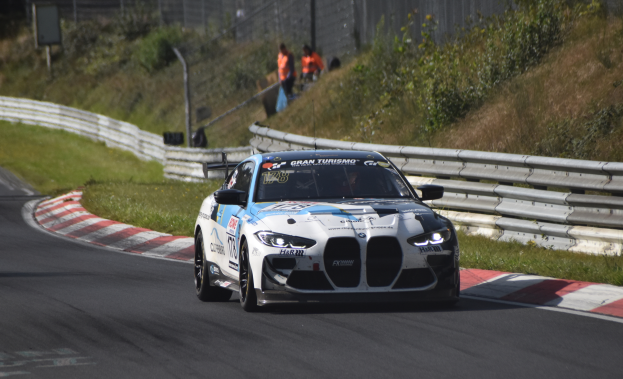 Ein BMW M8 GT3 Rennwagen fährt auf einer Rennstrecke, umgeben von einem Zaun, Gras, Pflanzen und einer Gruppe von Menschen, mit einem unscharfen Hintergrund.