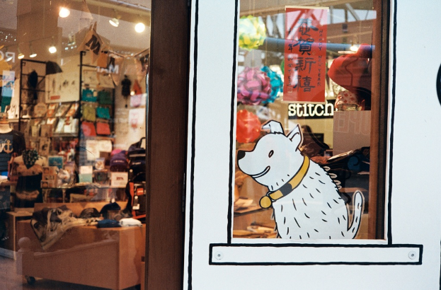 Eine Illustration eines Hundes mit Text auf der rechten Seite und links ein Raum mit Lampen, Fahnen, Regalen, Sofas, Kleidung, Dekorationen, Menschen, Stühlen und verschiedenen anderen Gegenständen.