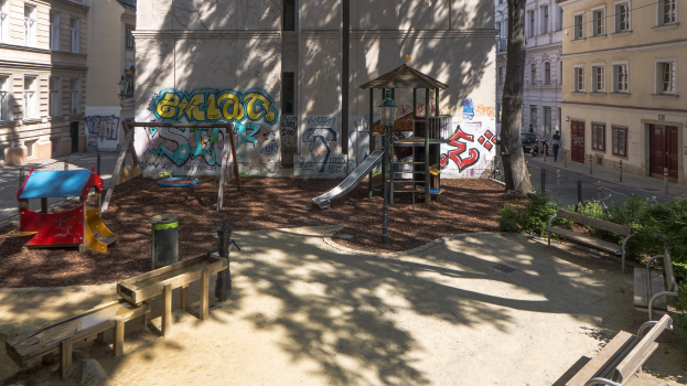 Ein Spielplatz in einer städtischen Umgebung mit Graffiti, umgeben von Gebäuden, Bäumen, Bänken, Pfählen und einem Mülleimer, mit Menschen und Fahrzeugen auf der Straße und dem Himmel im Hintergrund.