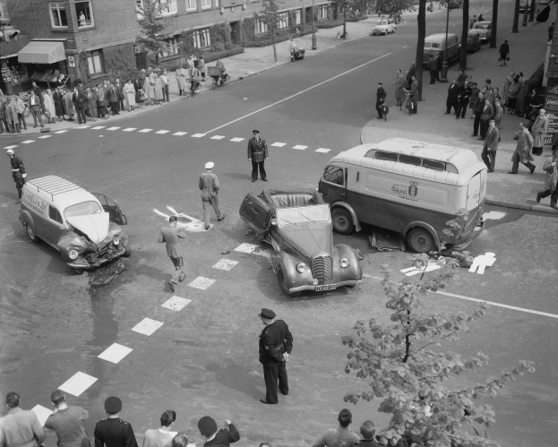 Schwarz-weiß-Szene eines Autounfalls an einer Straßenkreuzung mit mehreren Fahrzeugen, darunter ein Lieferwagen, und einer Gruppe von Schaulustigen; Gebäude, Bäume, Pfosten und Schilder im Hintergrund.