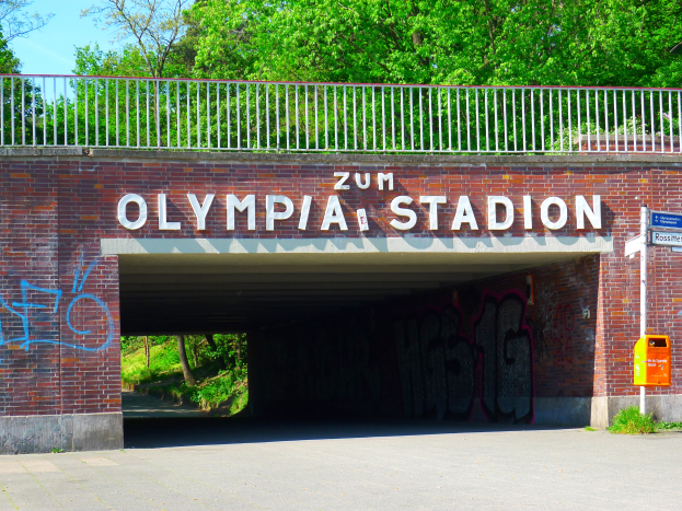 Der Eingang zum Olympiastadion in Berlin, Deutschland, mit einer Brücke, einem Metallzaun, einer Tafel, einem Kasten, Pflanzen, Gras, einer Baumgruppe und einem bewölkten Himmel.