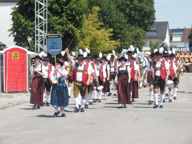 Eine Gruppe von Menschen in traditioneller bayerischer Tracht, einige mit Musikinstrumenten, geht eine Straße mit Bäumen, Gebäuden und einem klaren blauen Himmel im Hintergrund entlang, mit einem roten Objekt und einer Tafel auf der linken Seite.