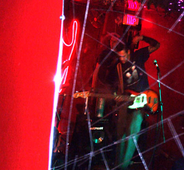 Ein Mann in einem schwarzen T-Shirt steht in der Mitte und hält eine Gitarre, während ein anderer Mann hinter ihm möglicherweise Instrumente spielt, unter hellen Scheinwerfern, vor einer roten Wand bei einem musikalischen Konzert.