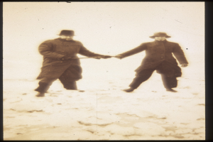 Zwei Männer in Mänteln und Hüten halten sich an den Händen und gehen durch unberührten Schnee, einer hält einen Hockey-Schläger.