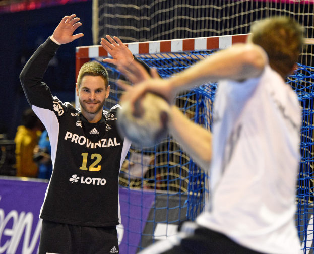 Zwei Männer spielen Handball vor einem Tor mit Netz und einem Banner mit Text im Hintergrund sowie einigen Zuschauern in einer Stadion.