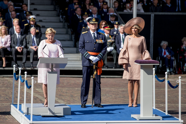 Eine Gruppe von Menschen auf einer Bühne im Königspalast in Amsterdam, mit einer Frau in einem rosa Kleid, die in ein Mikrofon spricht, und einem Mann mit einer Tasche, während andere sitzen und stehen.