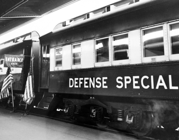 Ein Schwarz-Weiß-Foto eines stationären Zugs mit der Aufschrift 'Defense Special' und zwei Flaggen auf der linken Seite und einer Decke öberkopf.