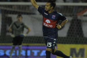 Ein Fußballspieler feiert auf dem Boden nach einem Tor, mit einer jubelnden Menge und einer Person im Hintergrund, alles vor einem unscharfen, energischen Hintergrund.