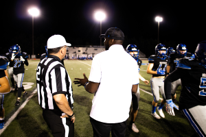 Ein Fußballtrainer mit Mütze spricht zu seinem Team auf einem Feld bei Nacht, mit Spielern in Helmen, Laternenmasten und einem dunklen Himmel im Hintergrund.