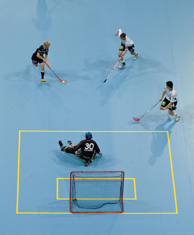 Drei Personen in Sportbekleidung spielen Hockey, wobei eine Person rechts ein Ball mit einem Hockeystock schlägt und eine Person hinter einem Netz auf dem Boden sitzt.