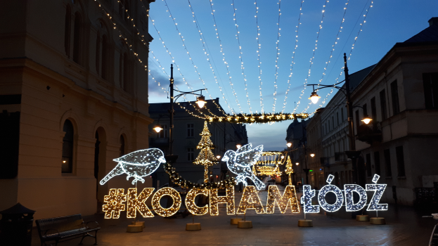 Eine belebte Straße in Krakau, Polen, geschmückt mit festlichen Weihnachtslichtern, gesäumt von Gebäuden, Straßenlaternen, Bänken und dekorativen Lichtern, unter einem sichtbaren Himmel.